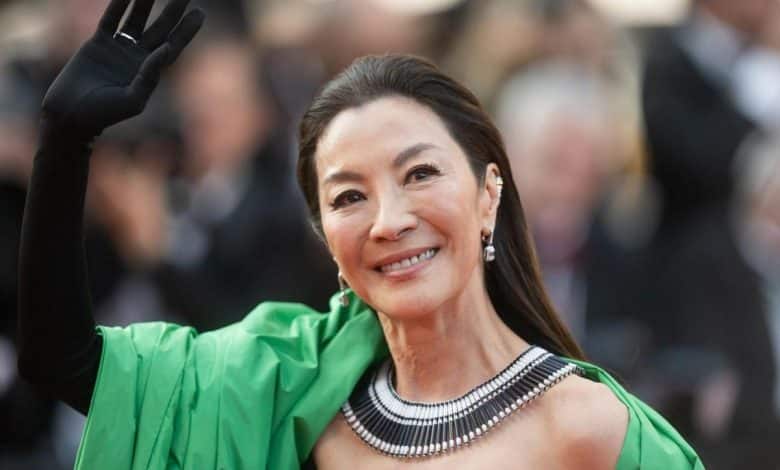 A woman in an elegant green dress waves at a camera on a red carpet, sporting a radiant smile. she has on a striking, multi-strand necklace and earrings, her long hair styled down.