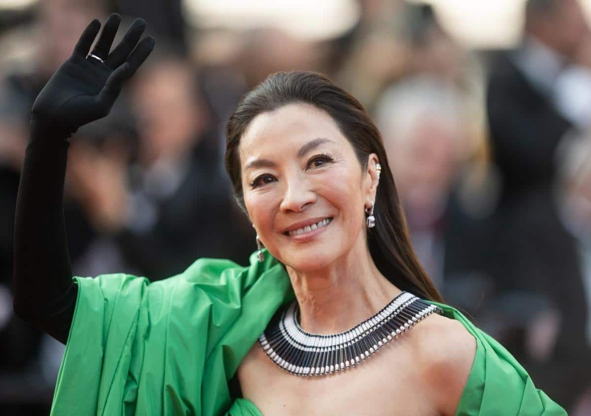A woman in an elegant green dress waves at a camera on a red carpet, sporting a radiant smile. she has on a striking, multi-strand necklace and earrings, her long hair styled down.