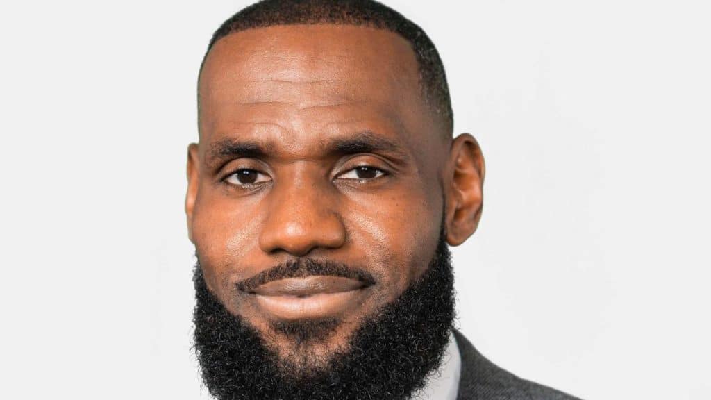 A bearded man with a close-cropped, neatly shaped black beard and a shaved head, smiling gently at the camera. he wears a gray suit, and is set against a simple, light-colored background.