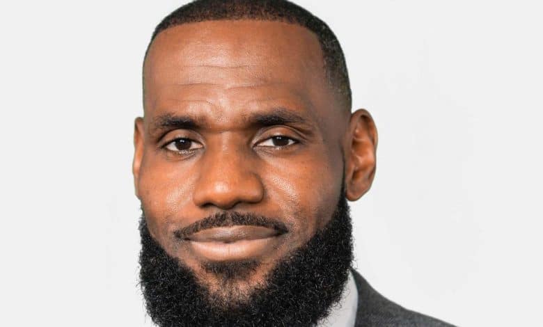 A bearded man with a close-cropped, neatly shaped black beard and a shaved head, smiling gently at the camera. he wears a gray suit, and is set against a simple, light-colored background.