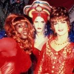 Three individuals in vibrant drag costumes pose together. on the left, one wears a red dress and blue feathers; in the middle, another sports a bold headdress and matching makeup; on the right, the last person is in a red lace outfit and a pearl necklace.