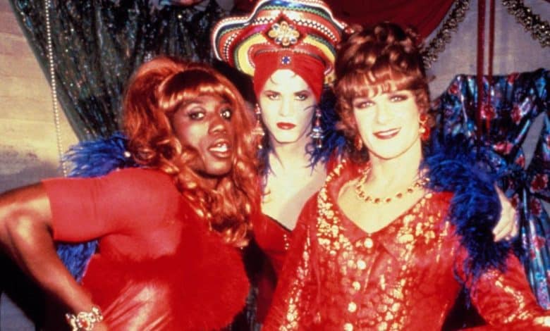 Three individuals in vibrant drag costumes pose together. on the left, one wears a red dress and blue feathers; in the middle, another sports a bold headdress and matching makeup; on the right, the last person is in a red lace outfit and a pearl necklace.