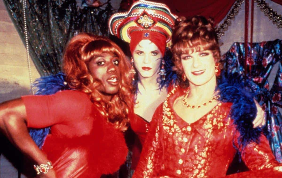 Three individuals in vibrant drag costumes pose together. on the left, one wears a red dress and blue feathers; in the middle, another sports a bold headdress and matching makeup; on the right, the last person is in a red lace outfit and a pearl necklace.