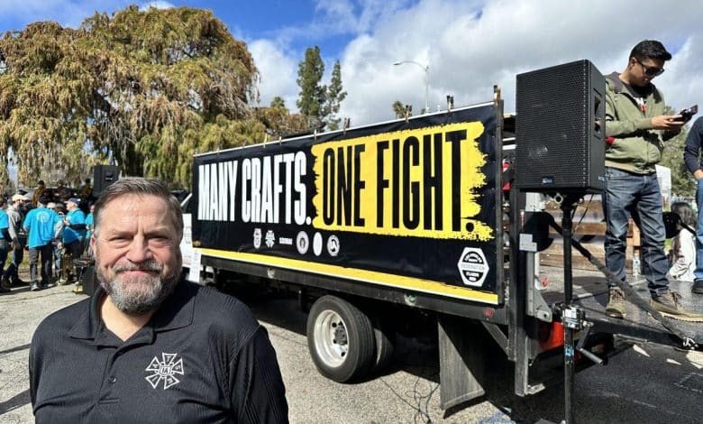 A man with a beard smiling in front of a trailer with the words "many crafts one fight" displayed. the trailer is equipped with speakers and there are people in the background under a cloudy sky.