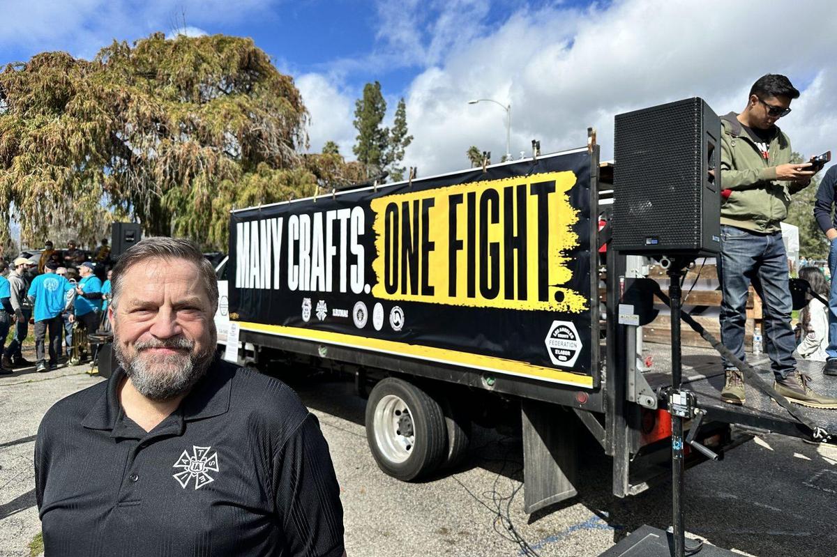 A man with a beard smiling in front of a trailer with the words "many crafts one fight" displayed. the trailer is equipped with speakers and there are people in the background under a cloudy sky.