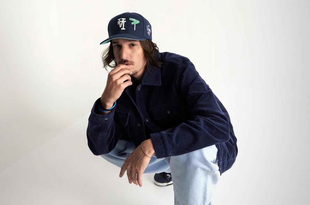 A young man crouches in a stylish pose, wearing a blue jacket, black pants, sneakers, and a dark cap with green embroidery. he has a thoughtful expression, touching his lips with one hand, against a plain white background.