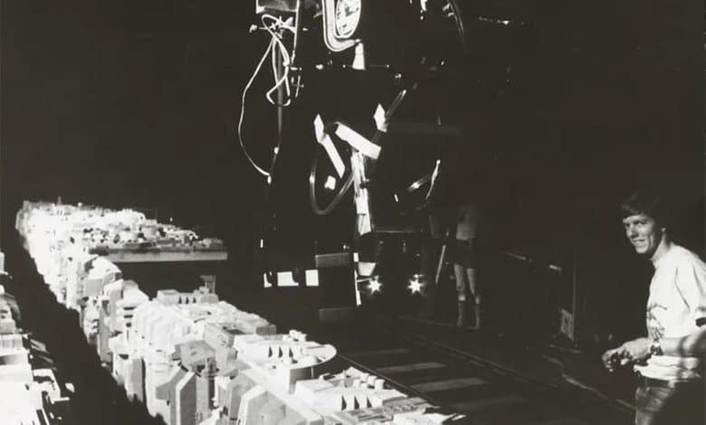 A black-and-white photo showing a woman controlling a robotic arm in a laboratory setting, surrounded by numerous neatly organized electronic components and tools on a long table, illuminated under focused lighting.