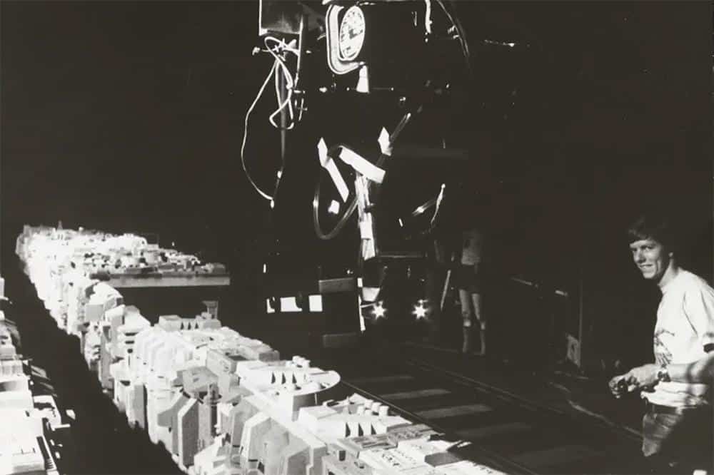 A black-and-white photo showing a woman controlling a robotic arm in a laboratory setting, surrounded by numerous neatly organized electronic components and tools on a long table, illuminated under focused lighting.