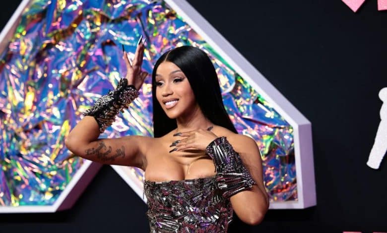 A woman poses on a red carpet, wearing a sparkly, strapless dress with off-shoulder details and intricate arm accessories. she has long, straight black hair and a bright smile, standing in front of a colorful, geometrically shaped backdrop.