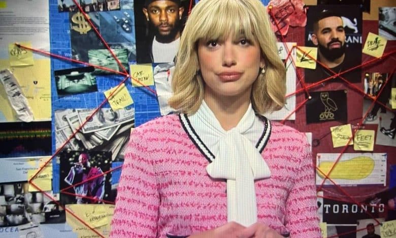 A woman with blonde hair and bangs wears a pink tweed jacket and white blouse with a tie. behind her is a corkboard covered in photos, notes, and maps, including images of celebrities and marked locations, creating a detective-style investigation board.