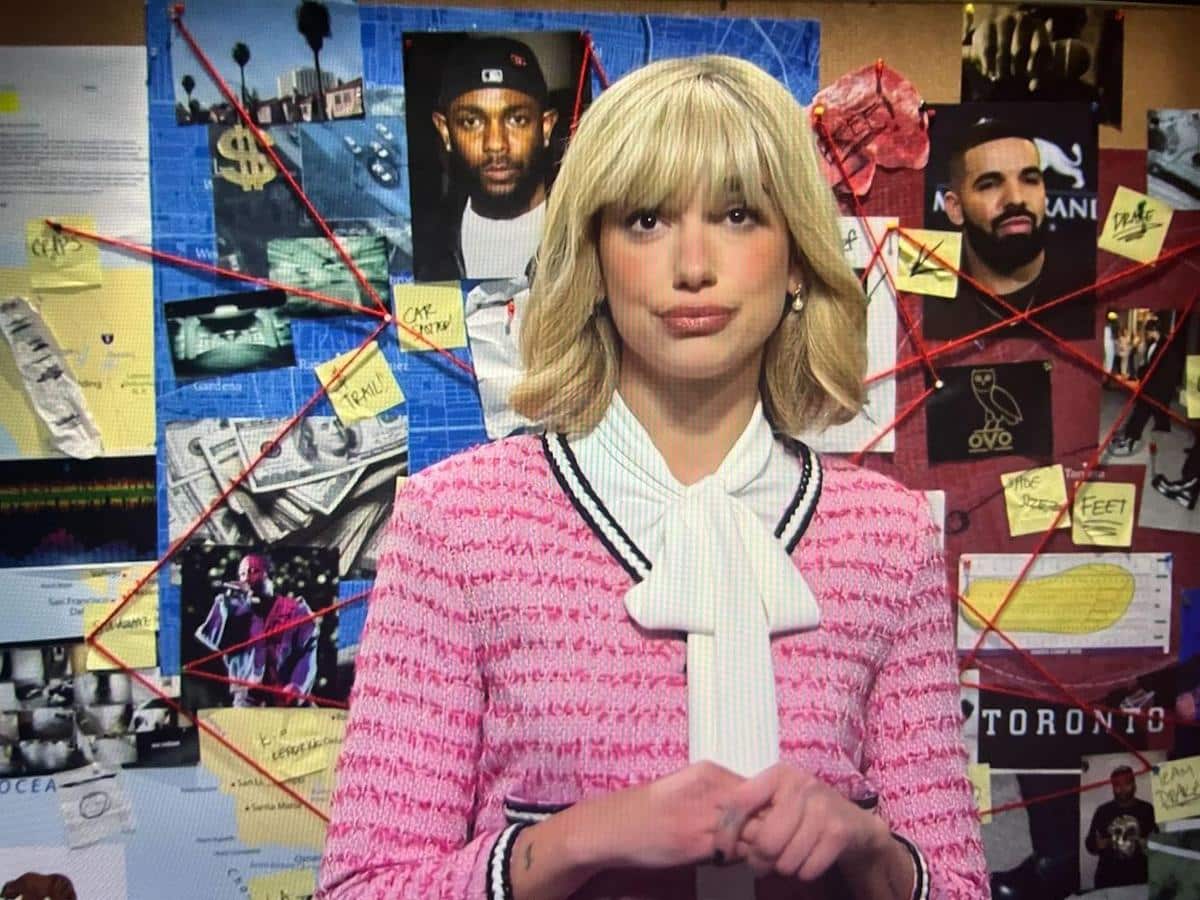 A woman with blonde hair and bangs wears a pink tweed jacket and white blouse with a tie. behind her is a corkboard covered in photos, notes, and maps, including images of celebrities and marked locations, creating a detective-style investigation board.