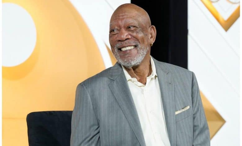 An elderly man with a beard smiles warmly, wearing a gray suit and cream shirt. he is seated, with a bright yellow circular design visible in the background.