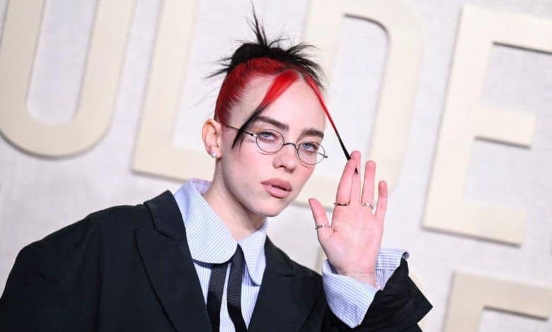A person with vibrant red and black hair styled upwards, wearing round glasses, a striped collar shirt, and a black jacket, gestures near their face while looking at the camera, standing against a pale background with blurred text.