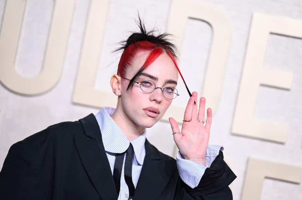 A person with vibrant red and black hair styled upwards, wearing round glasses, a striped collar shirt, and a black jacket, gestures near their face while looking at the camera, standing against a pale background with blurred text.