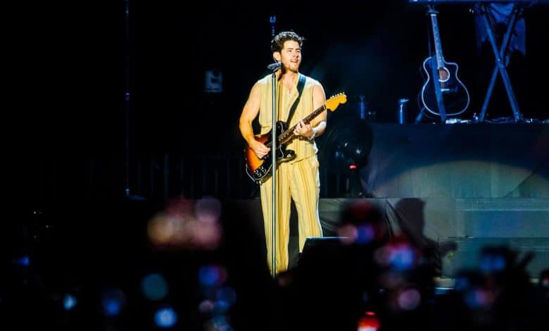 A musician plays an electric guitar on stage, wearing a flowing beige outfit. the stage is dimly lit, highlighted with blue hues, while the foreground shows the blurred heads of an audience in darkness, capturing the ambiance of a live concert.