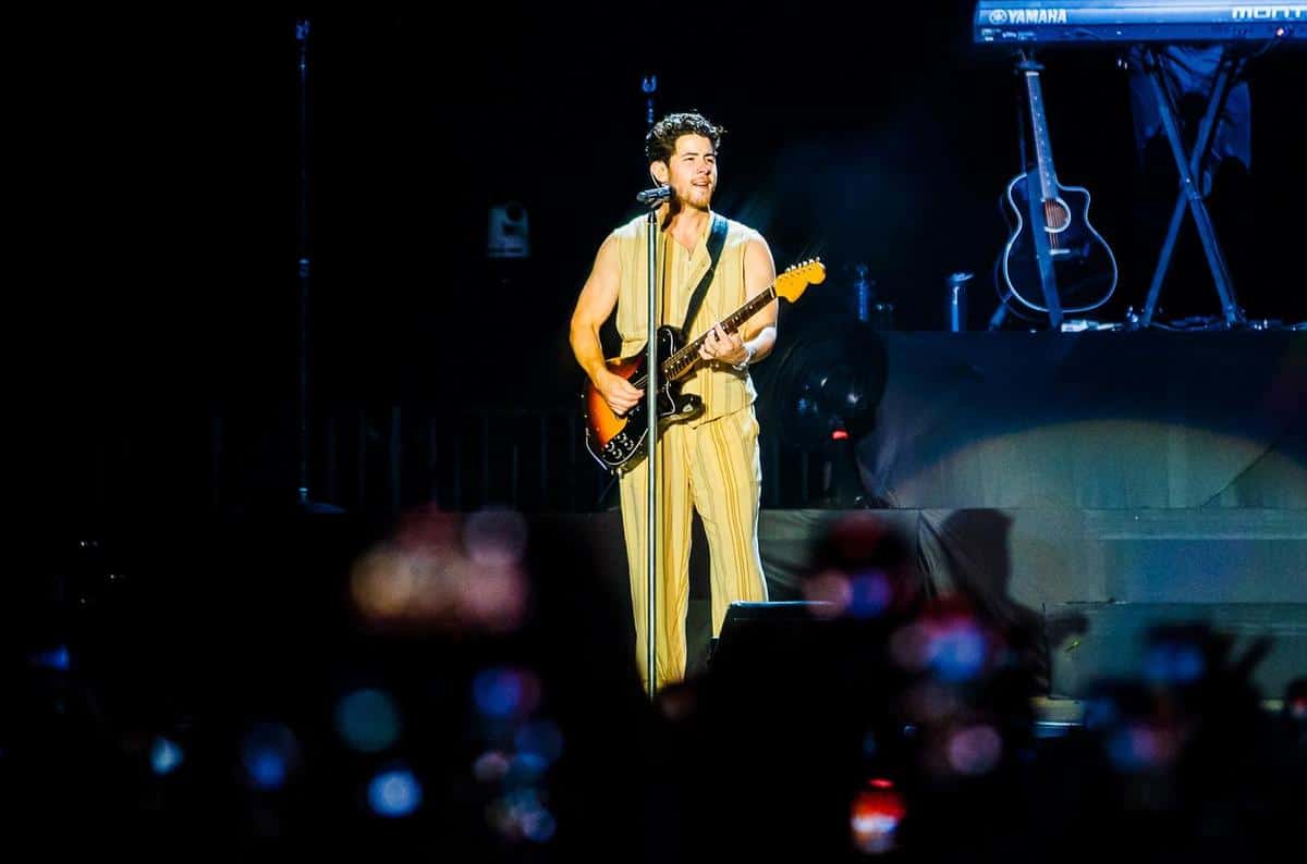 A musician plays an electric guitar on stage, wearing a flowing beige outfit. the stage is dimly lit, highlighted with blue hues, while the foreground shows the blurred heads of an audience in darkness, capturing the ambiance of a live concert.