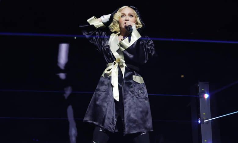 A female performer sings into a microphone on stage, wearing a black robe with yellow accents. she's surrounded by a dark setting with blue-lit accents and a minimalist modern design.