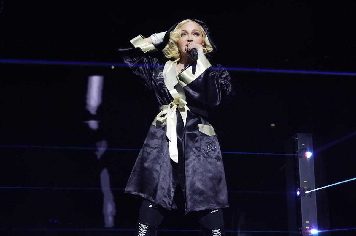 A female performer sings into a microphone on stage, wearing a black robe with yellow accents. she's surrounded by a dark setting with blue-lit accents and a minimalist modern design.