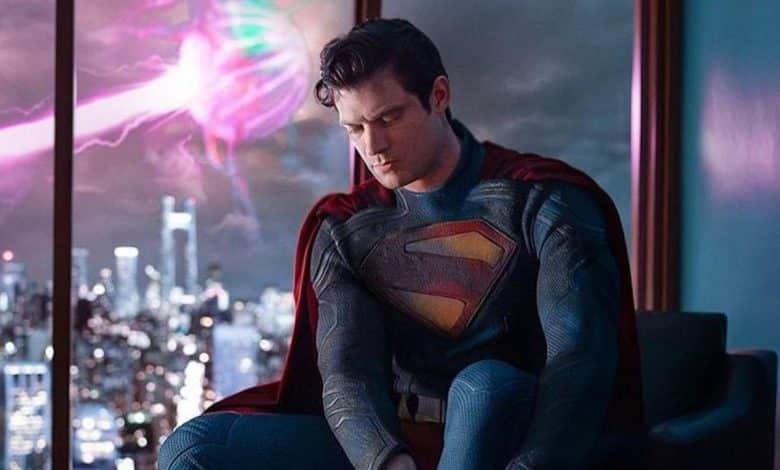 Superman, in his iconic blue and red suit with the 's' emblem, looks pensive while sitting near a window with a view of a city skyline at night, illuminated by a colorful aurora-like light in the sky.