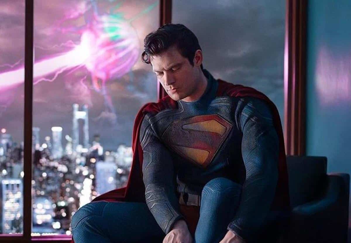 Superman, in his iconic blue and red suit with the 's' emblem, looks pensive while sitting near a window with a view of a city skyline at night, illuminated by a colorful aurora-like light in the sky.