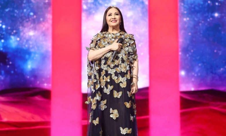 A woman stands onstage, wearing a black dress adorned with large metallic gold butterflies. she holds a microphone, smiling under bright stage lights with a vibrant red and pink background.