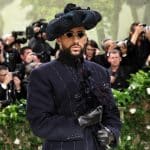 A man in a striking pinstripe suit and a large, ornate hat adorned with flowers, accessorized with dark gloves and sunglasses, poses at an event with photographers capturing the moment in the background.