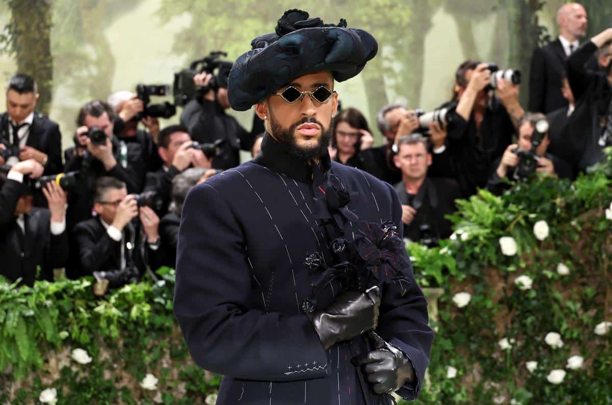 A man in a striking pinstripe suit and a large, ornate hat adorned with flowers, accessorized with dark gloves and sunglasses, poses at an event with photographers capturing the moment in the background.