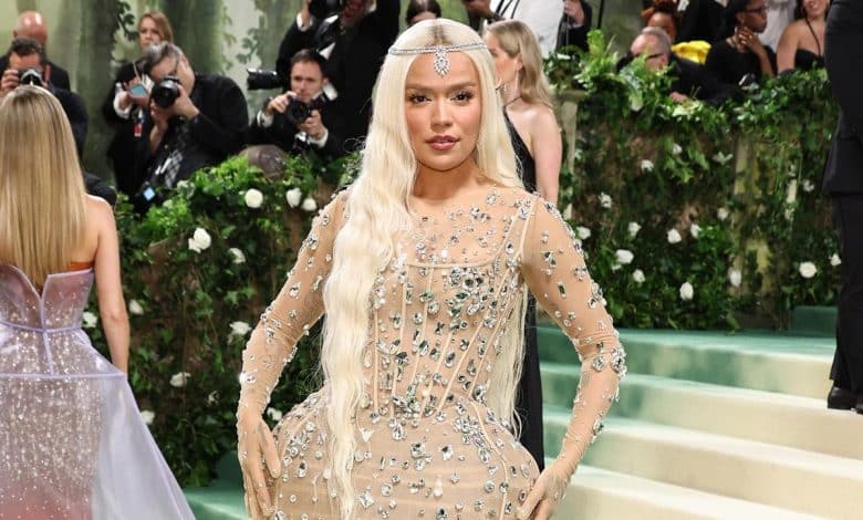 A woman at a glamorous event poses on a staircase, wearing a sheer, embellished gown with a headpiece. photographers capture the scene in the background, which is decorated with lush greenery and white flowers.