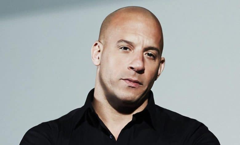 A bald man wearing a black shirt looks into the camera with a focused expression, set against a light gray background. his head is slightly tilted, and he has a serious facial expression.