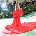 A woman in a striking red gown with a voluminous train stands on a stairway at a glamorous event, turning to wave, while photographers capture the moment. the background features a green staircase lined with white flowers.