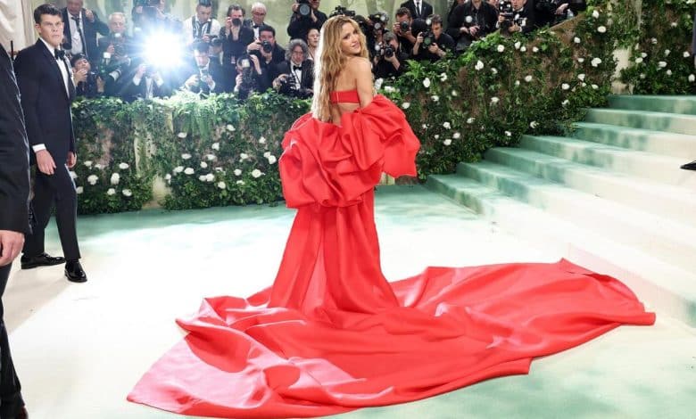 A woman in a striking red gown with a voluminous train stands on a stairway at a glamorous event, turning to wave, while photographers capture the moment. the background features a green staircase lined with white flowers.