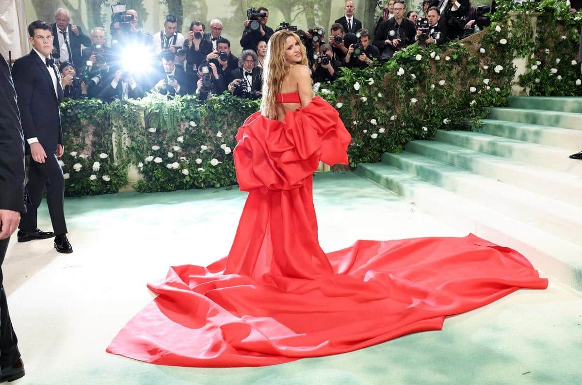 A woman in a striking red gown with a voluminous train stands on a stairway at a glamorous event, turning to wave, while photographers capture the moment. the background features a green staircase lined with white flowers.