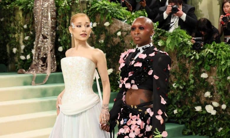 Two people posing on a staircase at a formal event. the person on the left wears a white, strapless gown with a pleated skirt, the other person is in a black outfit adorned with pink floral appliqués. photographers capture the moment in the background.