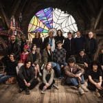 Group of diverse people posing together in a cozy attic room with a large stained glass window, decorative items hanging from the ceiling, and wooden interiors, conveying a warm, creative atmosphere.