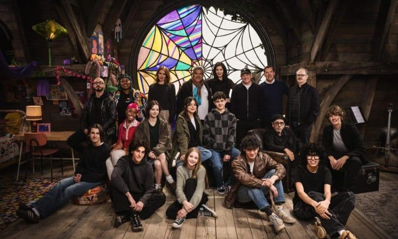 Group of diverse people posing together in a cozy attic room with a large stained glass window, decorative items hanging from the ceiling, and wooden interiors, conveying a warm, creative atmosphere.