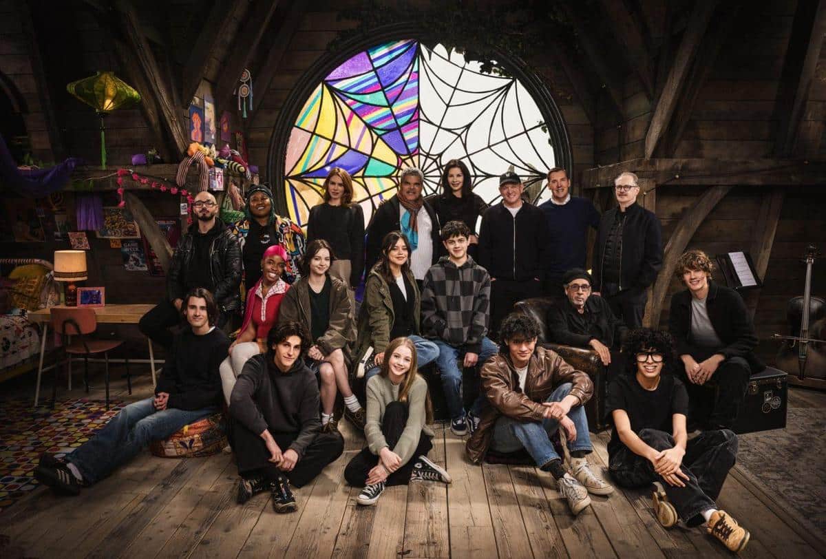 Group of diverse people posing together in a cozy attic room with a large stained glass window, decorative items hanging from the ceiling, and wooden interiors, conveying a warm, creative atmosphere.