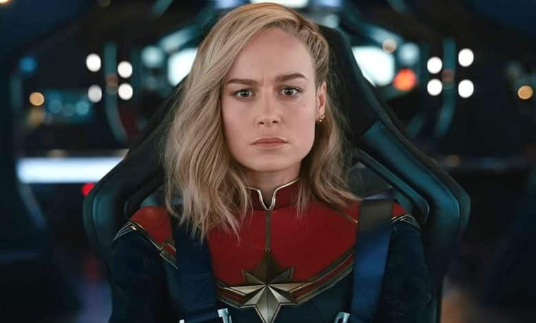 A woman in a captain marvel costume sits inside a futuristic cockpit, looking serious and determined. she has shoulder-length blonde hair and wears a blue, red, and gold suit with a prominent star emblem.