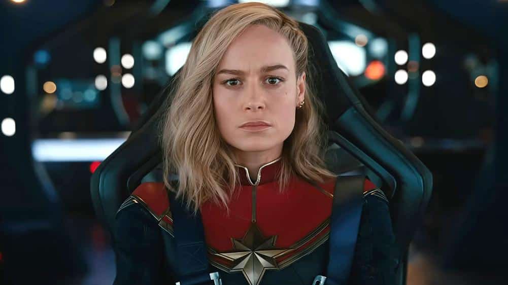 A woman in a captain marvel costume sits inside a futuristic cockpit, looking serious and determined. she has shoulder-length blonde hair and wears a blue, red, and gold suit with a prominent star emblem.