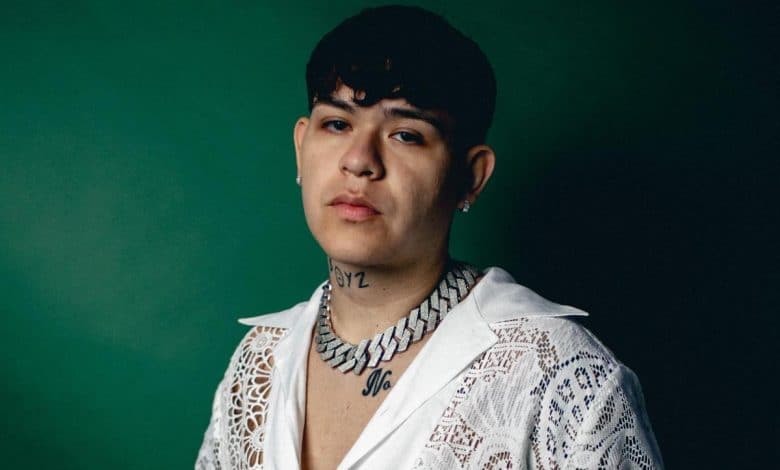 A young man with short dark hair and visible tattoos on his neck, wearing a white lace top, poses against a dark green background. his expression is serious and contemplative.