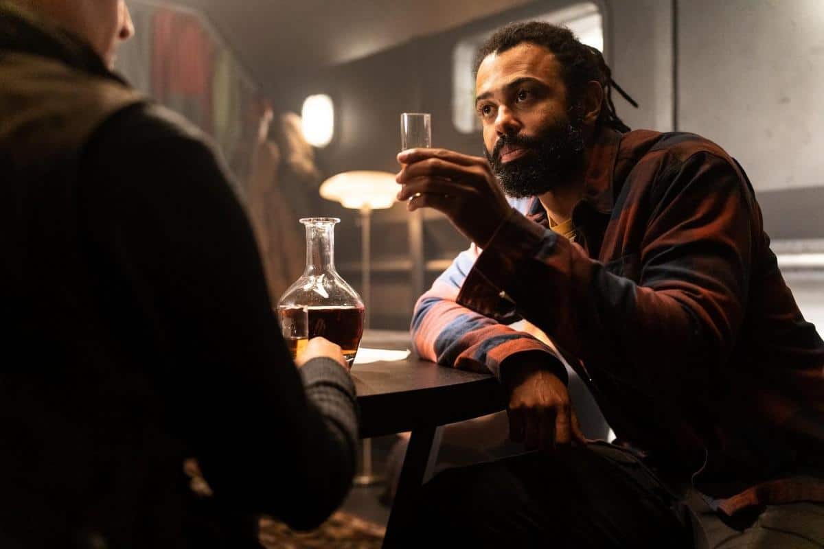 A man with a beard and dreadlocks sits at a table, holding a glass of liquor. he is engaged in a conversation in a dimly-lit bar, with a decanter and another person across the table partially visible.