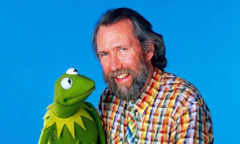 A man with a beard and plaid shirt smiling next to kermit the frog against a bright blue background. kermit appears animated with his big eyes and green body, looking at the camera.