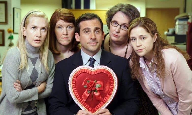 Five people pose in an office setting; four women and one man in the center holding a large heart-shaped box. the man, with a slight smile, wears a suit, and the women show varied expressions of surprise and curiosity.