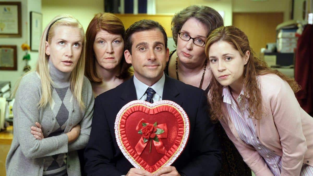 Five people pose in an office setting; four women and one man in the center holding a large heart-shaped box. the man, with a slight smile, wears a suit, and the women show varied expressions of surprise and curiosity.