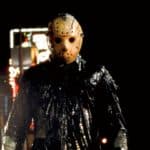 Jason voorhees in his iconic hockey mask and weathered clothing, stands on a rain-drenched street at night, illuminated by neon lights in the blurry background.