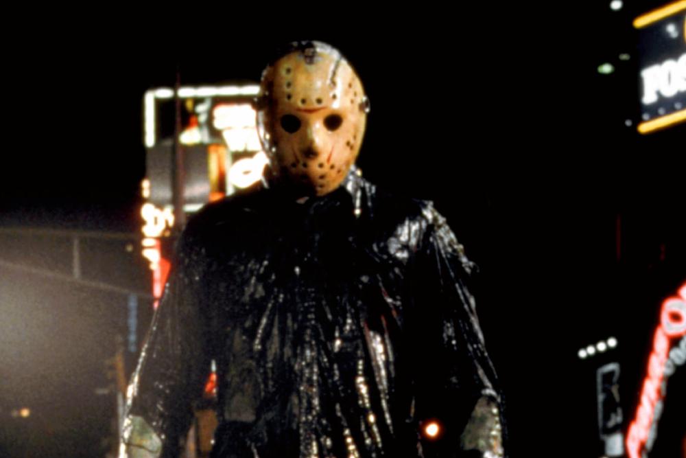 Jason voorhees in his iconic hockey mask and weathered clothing, stands on a rain-drenched street at night, illuminated by neon lights in the blurry background.