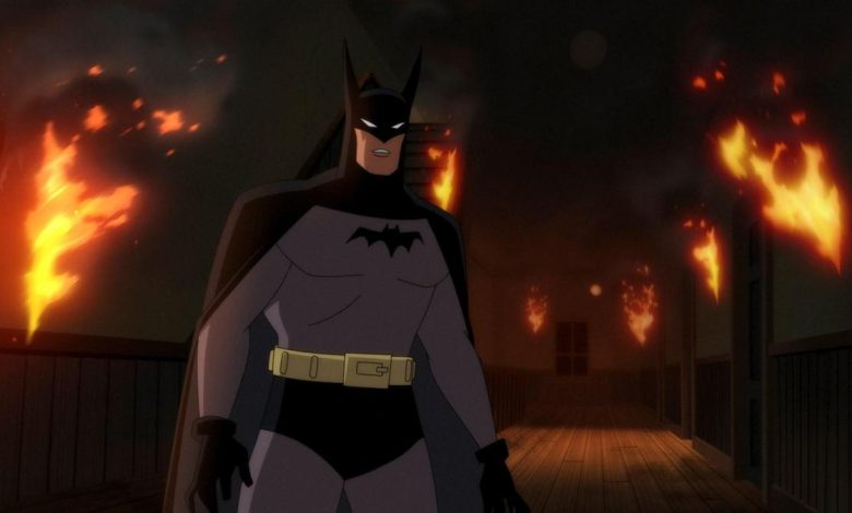 Batman stands on a wooden bridge at night, with a stern expression. behind him, buildings are engulfed in flames, illuminating the dark scene with fiery orange light.