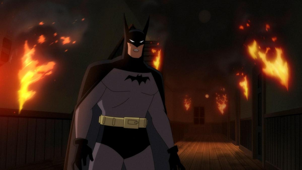 Batman stands on a wooden bridge at night, with a stern expression. behind him, buildings are engulfed in flames, illuminating the dark scene with fiery orange light.