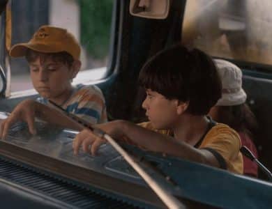 Two boys sit inside an old car, looking intently out the windshield. the boy on the left wears a yellow cap and a pensive expression, while the one on the right, in a yellow t-shirt, appears curious and focused.