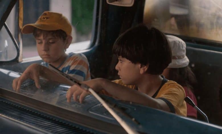 Two boys sit inside an old car, looking intently out the windshield. the boy on the left wears a yellow cap and a pensive expression, while the one on the right, in a yellow t-shirt, appears curious and focused.