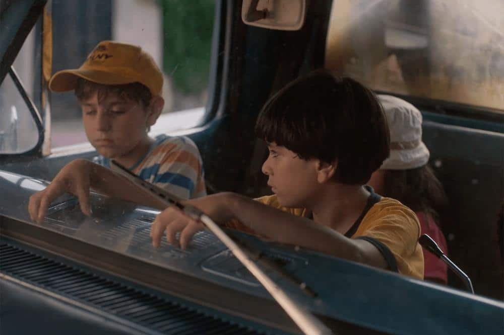 Two boys sit inside an old car, looking intently out the windshield. the boy on the left wears a yellow cap and a pensive expression, while the one on the right, in a yellow t-shirt, appears curious and focused.